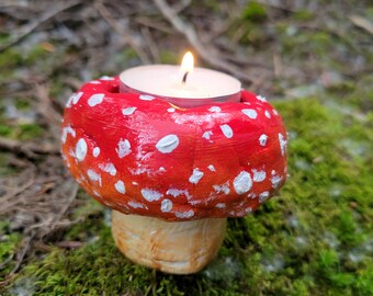 Può includere: Un portacandele in ceramica rosso e bianco a forma di fungo con una candela bianca al suo interno. Il portacandele è su un letto di muschio verde.