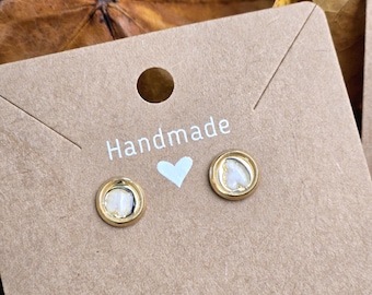 Può includere: Un paio di orecchini a bottone color oro con un design a forma di cuore. Gli orecchini sono posizionati su una carta marrone con la scritta "Handmade" stampata sopra un piccolo cuore blu. Gli orecchini hanno un centro bianco.