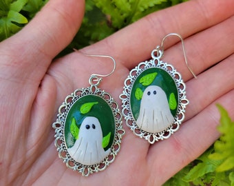 Può includere: Un paio di orecchini d'argento con ciondoli fantasma verdi e bianchi. I ciondoli sono incastonati in cornici ovali con un bordo decorativo.