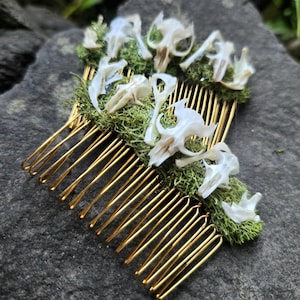 May include: A unique hair comb featuring gold-colored metal tines. The comb is adorned with small, white bone-like structures and green moss. The comb is set against a dark, textured background, creating a striking contrast.
