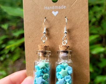 Può includere: Orecchini fatti a mano con piccole bottiglie di vetro riempite di stelle origami verde acqua e verde. Le bottiglie sono attaccate a ganci per orecchini argentati e sono esposte su una carta marrone con la scritta "Handmade".