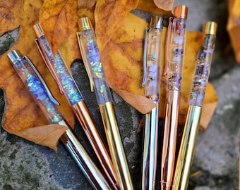 Può includere: Un assortimento di penne decorative con fusti trasparenti riempiti di glitter colorati e piccoli abbellimenti. Le penne hanno dettagli metallici in argento, oro e rame. Le penne sono disposte su foglie autunnali.