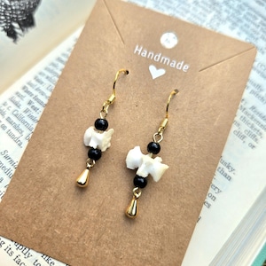 May include: Handmade earrings with a unique design. Each earring features a small, white bone-shaped element, black beads, and a gold-colored teardrop charm. The earrings hang from gold-colored hooks. The background includes a book and a brown card.