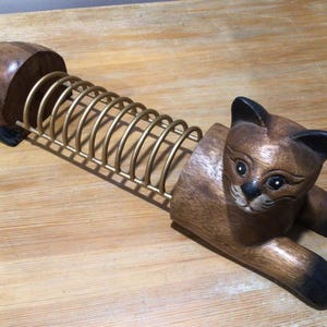 May include: A wooden cat-shaped letter rack with a brass-coloured spiral. The cat is carved from wood with brown and black accents. The letter rack is designed to hold mail or documents, adding a decorative touch to any desk or office.