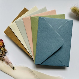 May include: A selection of coloured envelopes in shades of brown, cream, pink, green, and blue, fanned out on a white surface. A small bouquet of dried flowers and a cream-coloured ribbon are in the background. Perfect for stationery.