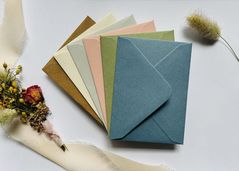 May include: A selection of coloured envelopes in shades of brown, cream, pink, green, and blue, fanned out on a white surface. A small bouquet of dried flowers and a cream-coloured ribbon are in the background. Perfect for stationery.