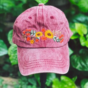 May include: A burgundy baseball cap with an embroidered floral design featuring yellow sunflowers, orange and red flowers, and green leaves.