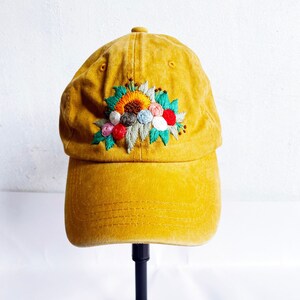 May include: A mustard yellow baseball cap with an embroidered floral design in shades of green, red, yellow, blue, and white.