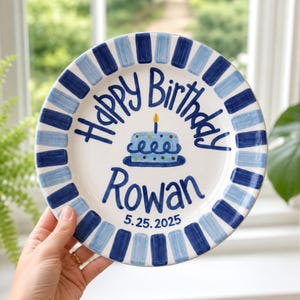 Pode incluir: Um prato redondo de cerâmica com centro branco e borda de listras azuis. O centro apresenta o texto Happy Birthday, uma ilustração de bolo azul com uma vela amarela, o nome Rowan e a data 25.05.2025 em letras azul-escuras.