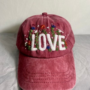 May include: A maroon baseball cap with the word "LOVE" embroidered in white with green leaves and colourful flowers surrounding the letters.