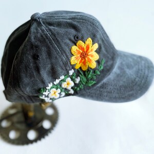May include: A dark grey baseball cap with embroidered yellow and white flowers. The cap has a curved brim and a mesh back.