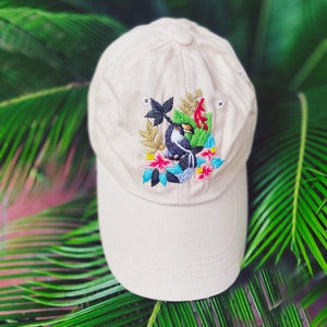 May include: A beige baseball cap featuring an embroidered toucan and tropical flowers. The toucan is black with a yellow beak and red accents. The flowers are pink, blue, and yellow.
