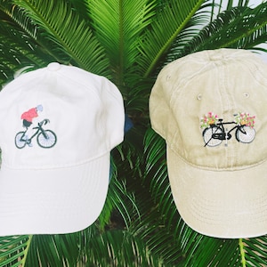Könnte beinhalten: Zwei Baseballkappen mit gestickten Designs. Eine Kappe ist weiß mit einem schwarzen Fahrrad und einer Person, die darauf fährt. Die andere Kappe ist beige mit einem schwarzen Fahrrad mit Blumen im Korb.