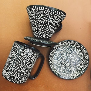 May include: A black ceramic coffee set with white floral patterns. The set includes a mug, a pour-over coffee maker, a saucer, and a matching mug. The designs are carved into the ceramic, creating a textured look. The set is displayed on a brown surface.