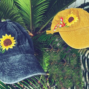 May include: Two embroidered baseball caps with floral designs. One cap is black with a sunflower and blue flowers, the other is yellow with a sunflower and red flowers.