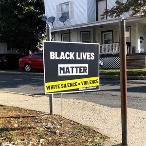 Może przedstawiać: Znak z napisem "BLACK LIVES MATTER" w białym tekście na czarnym tle, z żółtym banerem z napisem "WHITE SILENCE = VIOLENCE". Znak jest wyświetlany na zewnątrz na metalowym słupie przed dzielnicą mieszkalną.