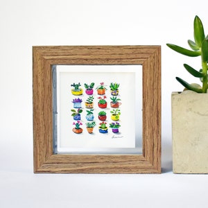 May include: A framed watercolor print featuring a collection of colorful potted plants. The frame is made of light brown wood. Next to the frame is a small concrete planter with a green succulent.