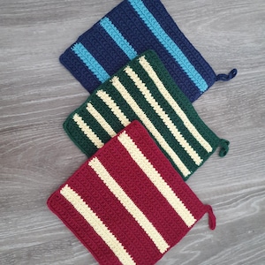 Op de afbeelding: Drie handgemaakte gehaakte pannenlappen in marineblauw, groen en bordeauxrood, elk met witte strepen. De vierkante pannenlappen hebben een kleine lus om op te hangen. Het haakwerk is zichtbaar en toont de textuur en het vakmanschap.