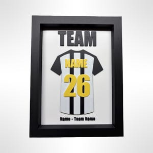 May include: Framed artwork featuring a black and white soccer jersey with the number 26 in yellow. The jersey has the word "NAME" in yellow and is set against a white background. The word "TEAM" is above the jersey, and "Name - Team Name" is below. The frame is black.