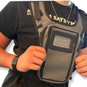 May include: A gray chest bag with black trim and a clear windowed pocket. The bag has adjustable straps and a zippered compartment. The person is wearing a black t-shirt and a black bracelet.