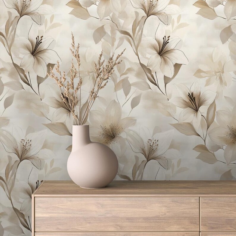 May include: A light pink vase with a rounded body holds dried grasses, set on a wooden surface. The background features a floral wallpaper design with large, stylized lilies in shades of beige and brown.