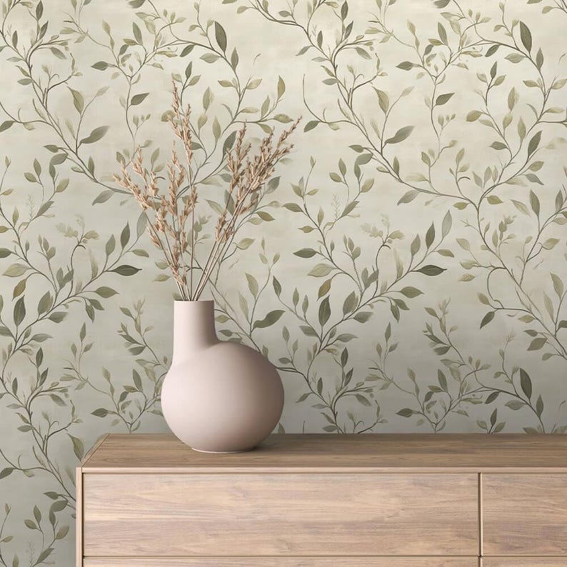 May include: A light pink vase holds dried ornamental grass, set on a wooden cabinet. The background features a wallpaper design with a repeating pattern of green and brown leaves and vines on a cream-colored backdrop. The overall aesthetic is natural and botanical.