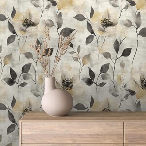 May include: A light pink ceramic vase with a rounded shape holds dried wheat stalks. The vase sits on a light wood dresser. The background is a floral wallpaper with cream, gray, and yellow flowers and leaves.