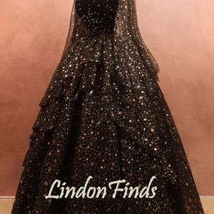 Könnte beinhalten: Ein schwarzes Abendkleid mit herzförmigem Ausschnitt und einem mehrschichtigen Rock. Das Kleid ist mit goldenen sternförmigen Pailletten verziert. Der Text "Lindon Finds" ist auf den Rock gedruckt.