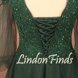 Op de afbeelding: Smaragdgroene avondjurk met een V-hals en lange, transparante mouwen. De bodice is versierd met glinsterende groene pailletten en een korsetrug. De tekst "Lindon Finds" is zichtbaar op de rok.