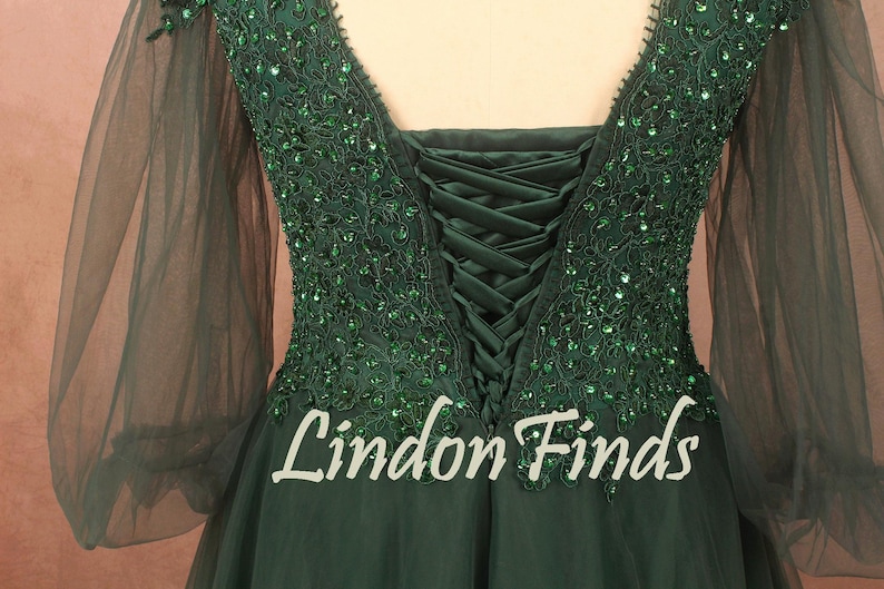 Op de afbeelding: Smaragdgroene avondjurk met een V-hals en lange, transparante mouwen. De bodice is versierd met glinsterende groene pailletten en een korsetrug. De tekst "Lindon Finds" is zichtbaar op de rok.