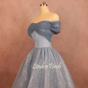 K&ouml;nnte beinhalten: Ein hellblaues, schulterfreies Kleid mit einem glitzernden T&uuml;llrock. Das Oberteil hat ein mehrlagiges T&uuml;lldesign und eine figurbetonte Silhouette. Das Kleid wird auf einer Schaufensterpuppe vor einem braunen Hintergrund gezeigt. Der Text "Lindon Finds" ist auf dem Rock sichtbar.