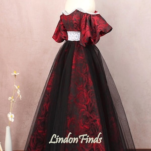 May include: A red and black off-the-shoulder gown with a floral print and a black tulle skirt. The dress has a white lace trim around the neckline and waist. The dress is on a mannequin and the background is a light pink.