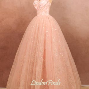May include: A pink tulle ball gown with a sweetheart neckline and floral embroidery. The dress has a fitted bodice and a full skirt. The skirt is adorned with sequins and a delicate floral pattern.