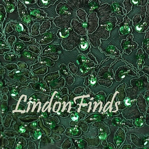 Op de afbeelding: Close-up van een donkergroene stof met ingewikkeld bloemenkant en glinsterende groene pailletten. De tekst "Lindon Finds" staat in een cursief lettertype over de stof. Het ontwerp is gedetailleerd en elegant.