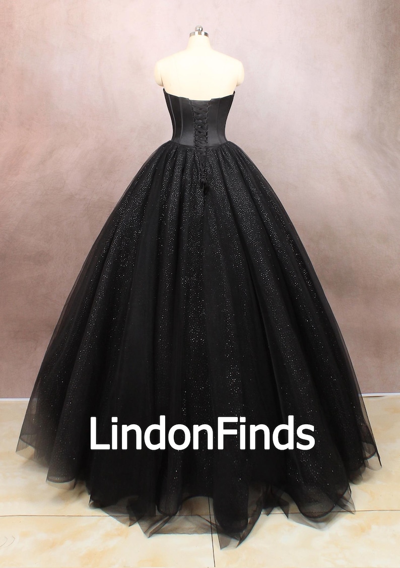 May include: A black strapless ball gown with a fitted bodice and a full skirt. The skirt is made of tulle and has a sparkly finish. The gown is cinched at the waist with a satin ribbon.