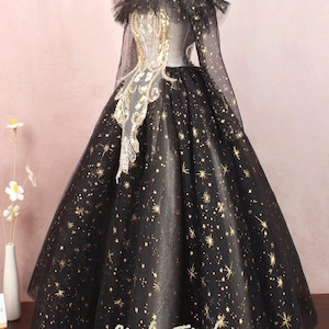May include: A black tulle ball gown with gold glitter stars. The gown has an off-the-shoulder neckline and long sleeves. The bodice is embellished with gold sequins and beads. The skirt is full and flowing. The gown is perfect for a special occasion.