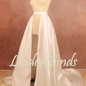 May include: A floor-length white satin overskirt featuring a high-waisted band and a dramatic front slit that reveals the interior. The skirt flows into a long train at the back, displayed on a beige mannequin. The text Lindon Finds is visible.