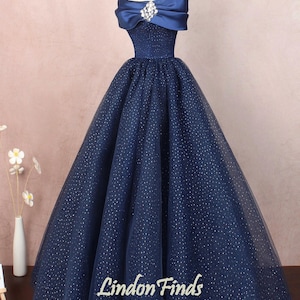 May include: A navy blue off-the-shoulder ball gown with a sparkly tulle skirt and a satin bodice. The dress has a bow detail at the neckline and a jeweled brooch.