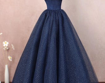 Op de afbeelding: Een marineblauwe off-shoulder baljurk met een glinsterende tule rok en een satijnen lijfje. De jurk heeft een strikdetail aan de halslijn en een broche met edelstenen.