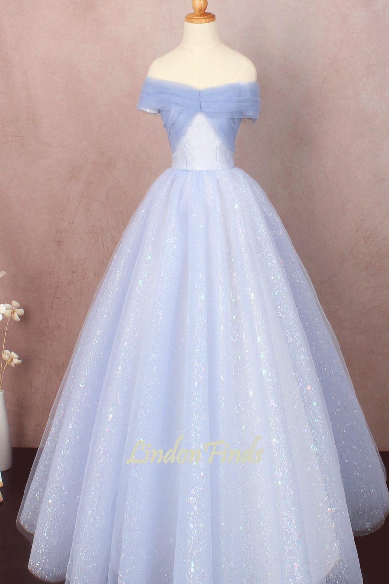 K&ouml;nnte beinhalten: Ein hellblaues T&uuml;ll-Ballkleid mit einem glitzernden, schillernden Finish. Das Kleid hat einen Off-Shoulder-Ausschnitt und ein tailliertes Oberteil.