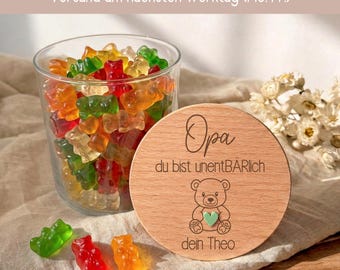 May include: A clear glass jar filled with multicolored gummy bears, accompanied by a round wooden lid engraved with the text 'Opa du bist unentBÄRlich' and 'dein Theo', featuring a small bear illustration and a tiny green heart.