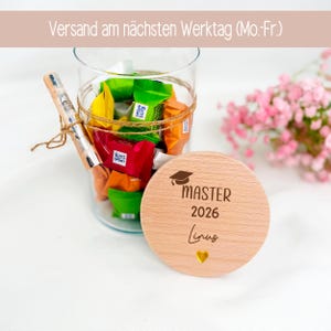 Könnte beinhalten: Ein Glasbehälter gefüllt mit bunten Süßigkeiten und einem Stift, der mit Schnur gebunden ist. Daneben ein Holzdeckel mit der Aufschrift "MASTER 2026 Linus" und einem Abschlusskappen-Symbol. Aufschrift "Versand am nächsten Werktag (Mo.-Fr.)".