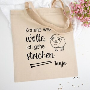 Könnte beinhalten: Eine beige Stofftasche mit dem Text "Komme was wolle, ich gehe stricken. Tanja" und einer schwarz-weißen Illustration eines Schafes. Die Tasche hat zwei schwarze Stricknadeln darauf gezeichnet.