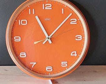 May include: Orange wall clock with white numerals and hands. The clock has a light brown border and a white power cord. The clock face is a bright orange color. The clock is on a wooden surface.