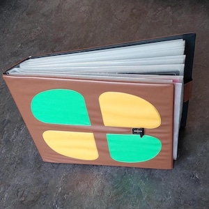 May include: A brown photo album with a four-leaf clover design in green and yellow. The album is closed with a black clasp and contains multiple clear sleeves for photos. The album is on a dark surface.