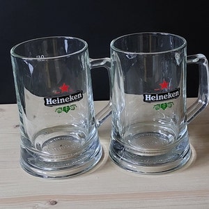 Può includere: Due boccali di birra in vetro trasparente con il logo Heineken e una stella rossa. I boccali hanno un manico sul lato destro e sono appoggiati su una superficie di legno.