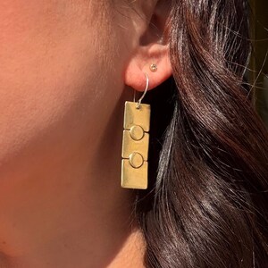 May include: Gold-toned rectangular earrings with a circular design. The earrings are composed of three stacked rectangles, each featuring a circle in the center. The earrings hang from silver hooks.