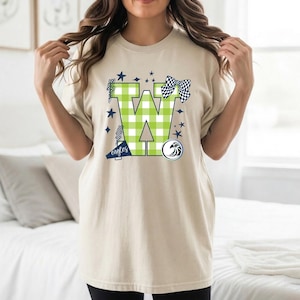 May include: Beige t-shirt with a large green and white checked "W" design, navy blue stars, a checkered bow, and a team logo. The word "eagles" is printed in navy blue. The shirt has a relaxed, oversized fit.