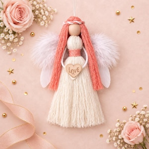 May include: A handmade angel ornament crafted from cream and coral-colored cord, featuring a smooth wooden bead head, fluffy white feather wings, and a small wooden heart pendant engraved with the word LOVE centered on the torso.