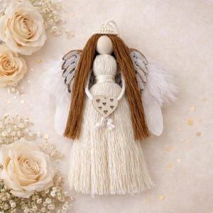May include: A handcrafted angel ornament made of cream-colored yarn with long brown yarn hair, a wooden bead head, and white feathery wings. A small wooden heart cutout with six tiny heart-shaped holes is suspended in front of the body.